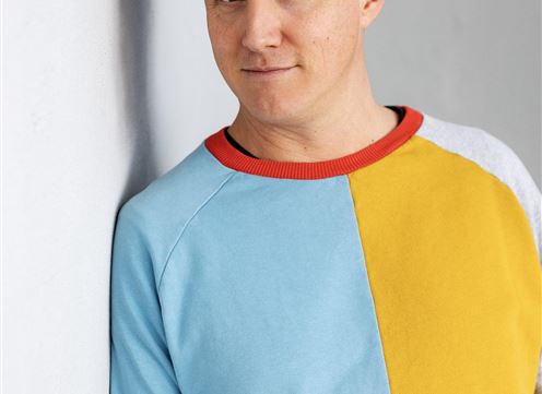 A man is standing by a wall and smiling. He is wearing a colorful sweater and a black beanie.