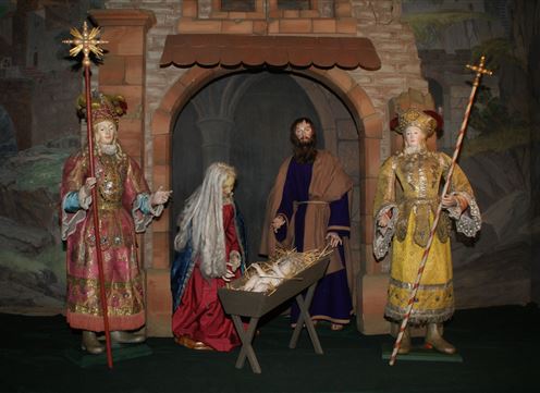 A nativity scene with figures representing the Holy Family. They stand in an architectural backdrop with a couch on which baby Jesus lies.