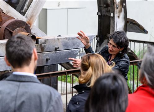 Een vrouw legt iets uit in de open lucht en wijst naar een grote machinestructuur. Een groep mensen luistert aandachtig.