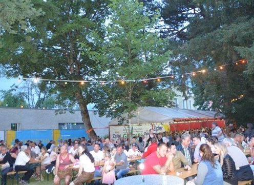 A lively outdoor event with many people sitting at tables and eating. The surroundings are decorated with lights, and there are trees visible in the background.