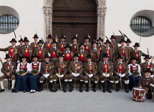 Un gruppo di persone in abbigliamento tradizionale si trova davanti a una grande porta. Indossano cappelli e medaglie e tengono in mano alcuni premi.