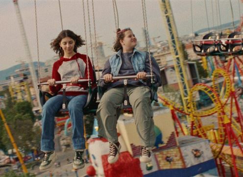 A man in a black suit stands on the stairs while a woman in an elegant pink dress sits. The room is modern and bright with a minimalist design.