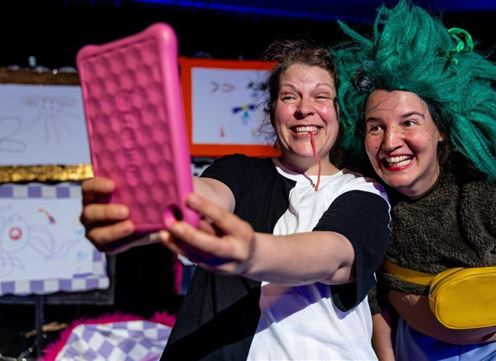 Two cheerful women are taking a selfie on a stage. Colorful drawings are visible in the background.