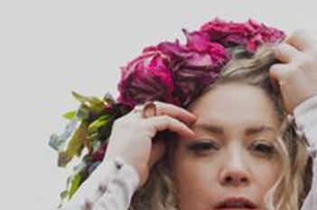 A woman with curly hair is wearing a wreath made of pink flowers. Her facial expression is thoughtful and dreamy.