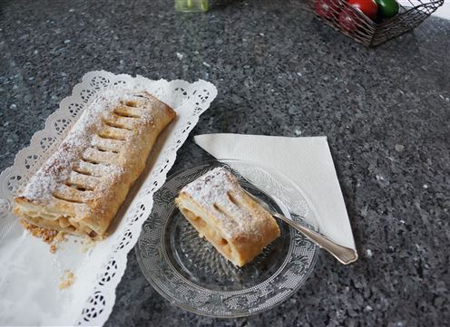Ein frisch gebackener Apfelstrudel auf einem weißen Teller. Daneben liegt ein Stück Strudel auf einem Glasteller.