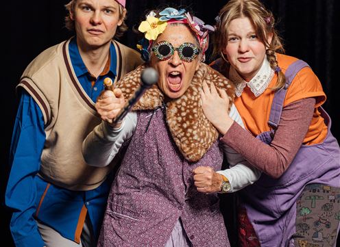 Drei Personen mit humorvollen Outfits posieren fröhlich. Die mittlere Person trägt eine auffällige Brille und hat einen energischen Ausdruck.