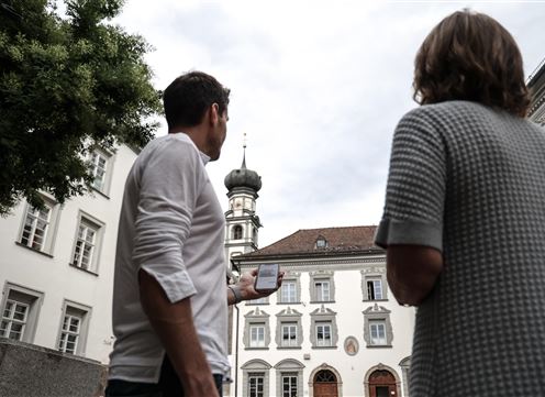 Two people are standing in a historical city view and looking at the tower of a building. The sky is cloudy and the atmosphere is calm.