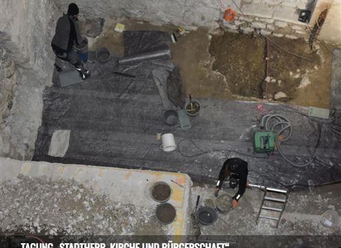 Eine Baustelle mit arbeitenden Personen in einem Kellerraum. Der Boden ist uneben und es sind verschiedene Werkzeuge und Materialien sichtbar.