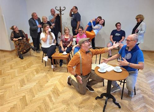 Eine Gruppe von Menschen sitzt und steht in einem Raum, einige lachen und interagieren miteinander. Im Vordergrund führen zwei Männer ein lustiges Gespräch an einem Tisch.