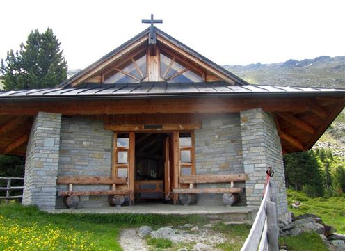 Eine kleine Kapelle aus Stein mit einem Kreuz auf dem Dach. Sie steht inmitten von grünen Wiesen und Bergen.
