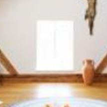 Ein heller Raum mit schrägen Holzdecken und einem großen Fenster. In der Mitte liegt ein runder Teppich, umgeben von Sitzkissen und Stühlen.