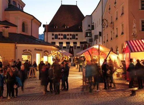 A bustling marketplace at dusk with colorful stalls and many people. The buildings in the background give the scene a charming, historic character.