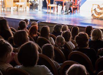 Un public est assis sur des chaises dans une salle et regarde une performance sur scène. Les spectateurs sont attentifs et profitent de la représentation.