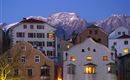 A charming city view with historic buildings and illuminated windows. Snow-capped mountains are visible in the background.