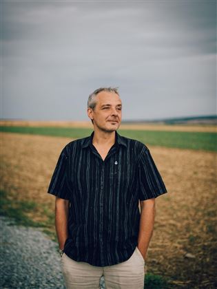 Ein Mann steht auf einem Feld, mit einer neutralen Miene. Der Himmel ist bewölkt und die Umgebung wirkt ländlich.