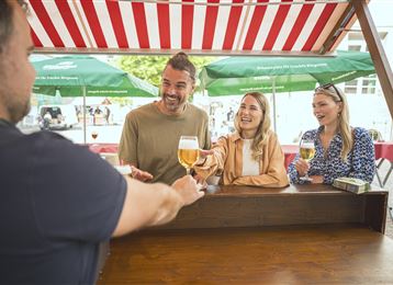 Eine fröhliche Gruppe von drei Personen steht an einem Stehtisch und genießt Getränke. Im Hintergrund sind grüne Sonnenschirme und ein festliches Ambiente zu sehen.