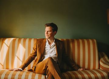 A man in a brown suit is sitting relaxed on a striped sofa. He is looking thoughtfully out of a window in a stylish room.