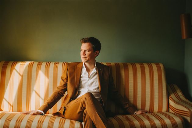 A man in a brown suit is sitting relaxed on a striped sofa. He is looking thoughtfully out of a window in a stylish room.