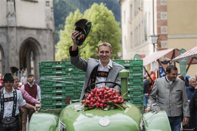Ein junger Mann fährt auf einem alten Traktor und trägt einen Hut. Er lächelt und winkt, während andere Menschen in traditioneller Kleidung im Hintergrund stehen.