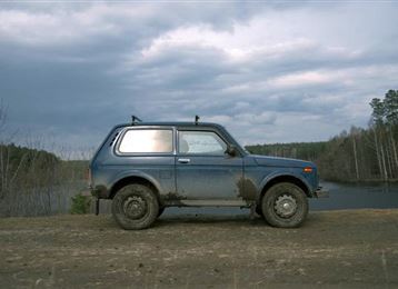 A blue car is parked on a narrow path next to a body of water. In the background, trees and a cloudy sky can be seen.