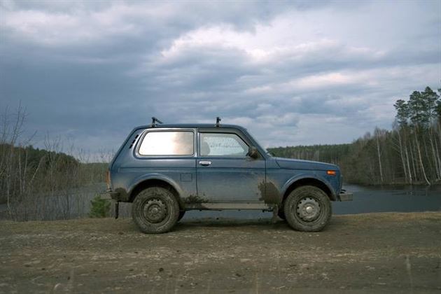 Une voiture bleue est garée sur un chemin étroit à côté d'un plan d'eau. À l'arrière-plan, on peut voir des arbres et un ciel nuageux.