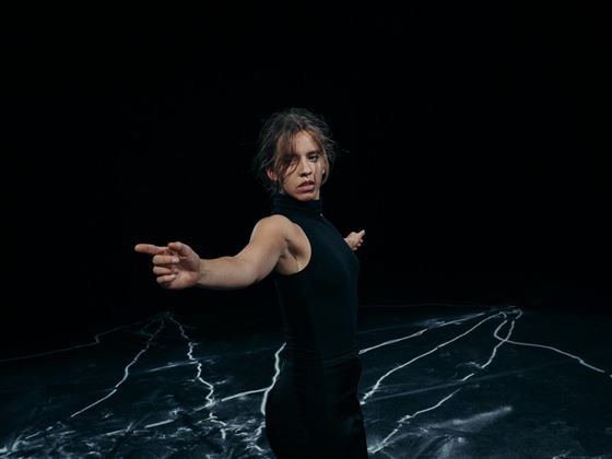 Une femme en vêtements sombres pointe du doigt dans une direction. L'arrière-plan est noir, et le sol a un design blanc avec des motifs.