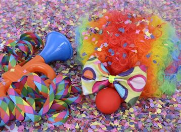 Des confettis colorés est éparpillé sur le sol, entouré d'une perruque rouge, d'un nœud coloré, d'un nez de clown rouge et d'un écureuil en jouet. Cela dégage une ambiance joyeuse de carnaval ou de fête.