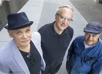 Tre uomini sono in piedi sul marciapiede e guardano verso la telecamera. Indossano diversi cappelli e stili di abbigliamento, mentre lo sfondo mostra un ambiente urbano.