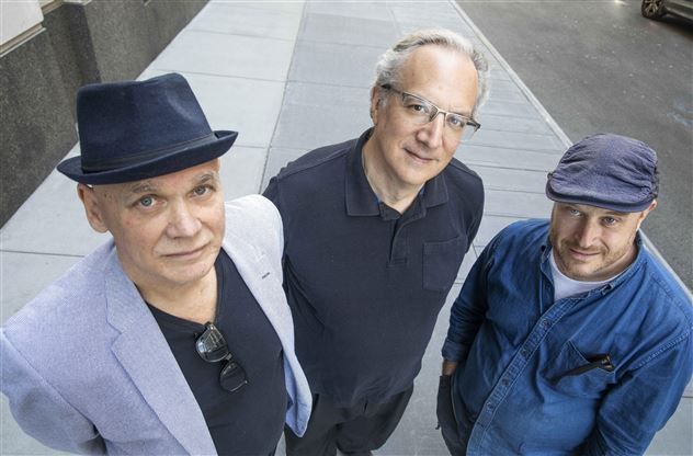 Three men are standing on the sidewalk and looking at the camera. They are wearing different hats and clothing styles, with the background showing an urban environment.