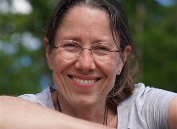 Eine lächelnde Frau mit Brille sitzt und stützt sich auf ihren Arm. Der Hintergrund ist grün und verwunschen.