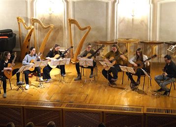 Un ensemble de musique se compose de sept guitaristes et d'un autre musicien sur scène. Ils jouent ensemble dans une salle de musique festive avec une ambiance chaleureuse.