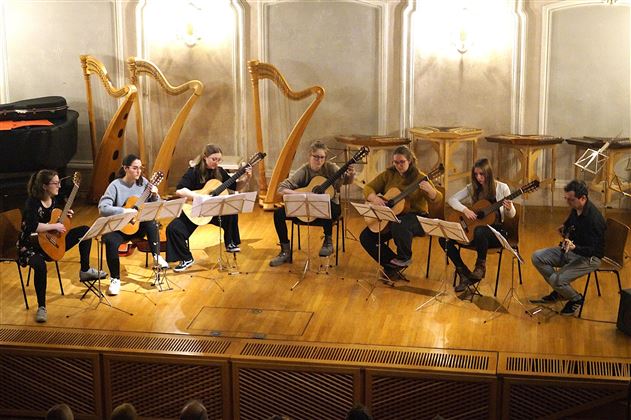 Ein Musikensemble besteht aus sieben Gitarristen und einem weiteren Musiker auf der Bühne. Sie spielen gemeinsam in einem festlichen Musiksaal mit einem warmen Ambiente.