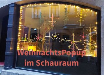 Ein hübsches Schaufenster mit festlicher Weihnachtsdekoration und bunten Lichtern. Der Schriftzug "WeihnachtsPopup im Schaurraum" zieht die Aufmerksamkeit auf sich.