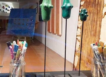 Eine künstlerische Ausstellung mit drei grünen Skulpturen auf Ständern. Im Vordergrund stehen Pinsel in einem Glas.