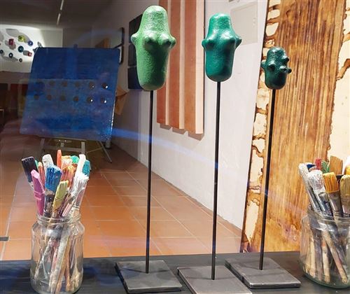Eine künstlerische Ausstellung mit drei grünen Skulpturen auf Ständern. Im Vordergrund stehen Pinsel in einem Glas.