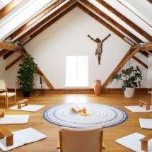 A bright room with sloping wooden ceilings and a large window. In the center lies a round rug, surrounded by cushions and chairs.