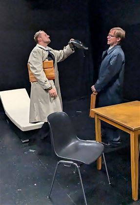 Deux hommes en costume se tiennent dans une pièce sombre. Le premier homme tient une chaussure et semble pensif, tandis que l'autre regarde attentivement.