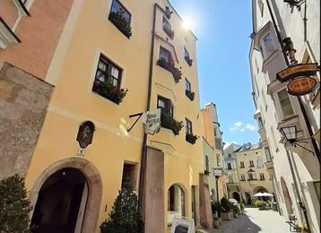 Eine malerische Gasse mit bunten Häusern und Blumenfensterbänken. Die Sonne scheint hell und hebt die charmante Atmosphäre des Ortes hervor.