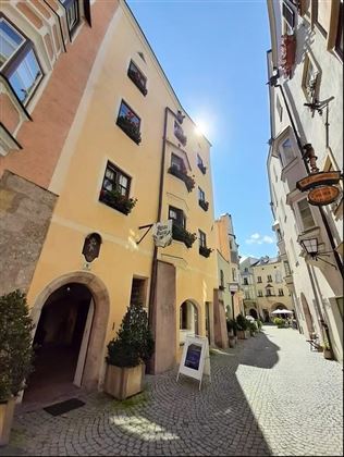 Eine malerische Gasse mit bunten Häusern und Blumenfensterbänken. Die Sonne scheint hell und hebt die charmante Atmosphäre des Ortes hervor.