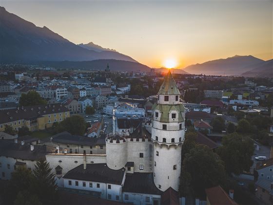 A historic tower with a green dome in front of a picturesque sunset. The city and the mountains in the background complete the landscape.