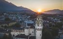A historic tower with a green dome in front of a picturesque sunset. The city and the mountains in the background complete the landscape.