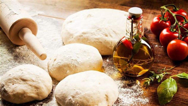 Fresh pizza dough lies on a wooden surface, surrounded by tomatoes and a bottle of olive oil. A rolling pin showcases the culinary style.