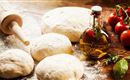 Fresh pizza dough lies on a wooden surface, surrounded by tomatoes and a bottle of olive oil. A rolling pin showcases the culinary style.