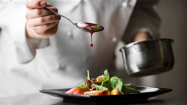 A chef decorates a salad with a sauce. The dish is presented on a black plate.