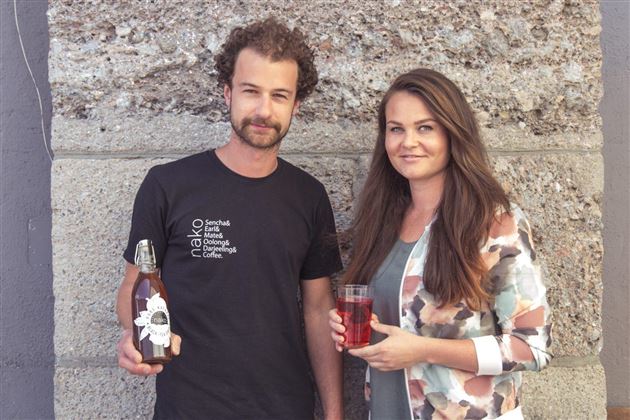 Deux personnes se tiennent devant un mur en pierre. L'homme tient une bouteille de boisson, tandis que la femme tient un verre avec un liquide rouge dans la main.