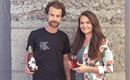 Deux personnes se tiennent devant un mur en pierre. L'homme tient une bouteille de boisson, tandis que la femme tient un verre avec un liquide rouge dans la main.