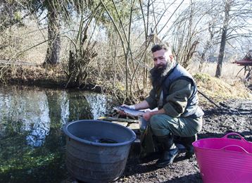 Un homme avec une barbe se tient au bord d'un petit plan d'eau et tient un poisson dans la main. À côté de lui, il y a deux récipients, un en plastique noir et un en rose.