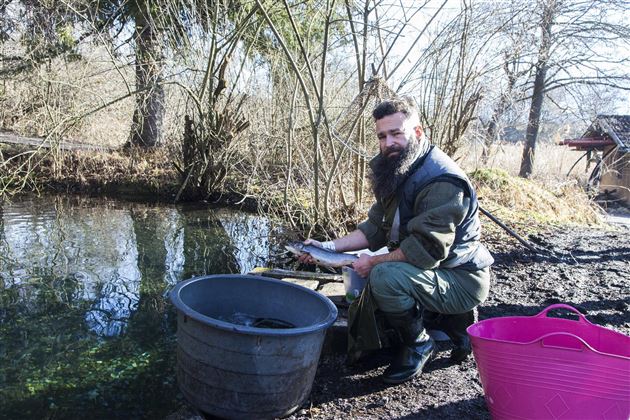 Een man met een baard staat aan de oever van een klein water en houdt een vis in zijn hand. Naast hem staan twee containers, een van kunststof in zwart en een in roze.