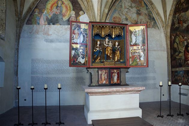 A historic altar with an open shrine displaying religious figures. In the background, walls with old frescoes and candle holders can be seen.