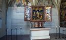 A historic altar with an open shrine displaying religious figures. In the background, walls with old frescoes and candle holders can be seen.